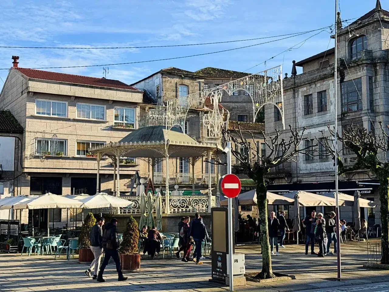 Gente paseando y disfrutando del sol en una plaza con terrazas, ideal para comer en Tui. Un quiosco y edificios históricos completan la escena.