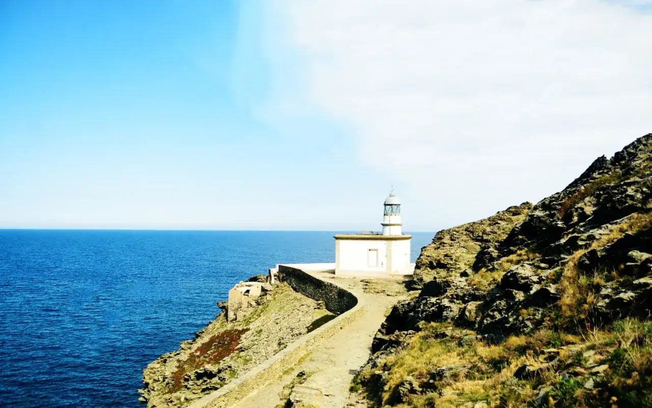 Un faro blanco se alza sobre un acantilado rocoso en la Costa Brava, con el mar azul extendiéndose hasta el horizonte.