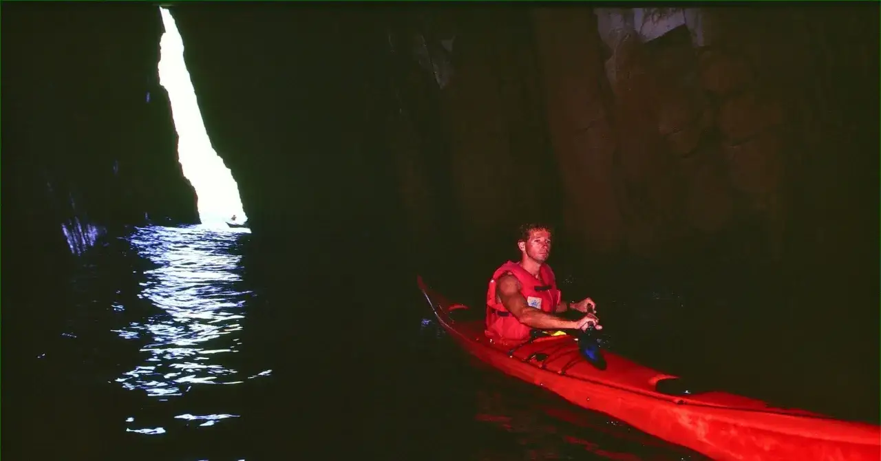Hombre en kayak rojo explora la Cova d'en Gispert, navegando por aguas oscuras hacia una abertura de luz.