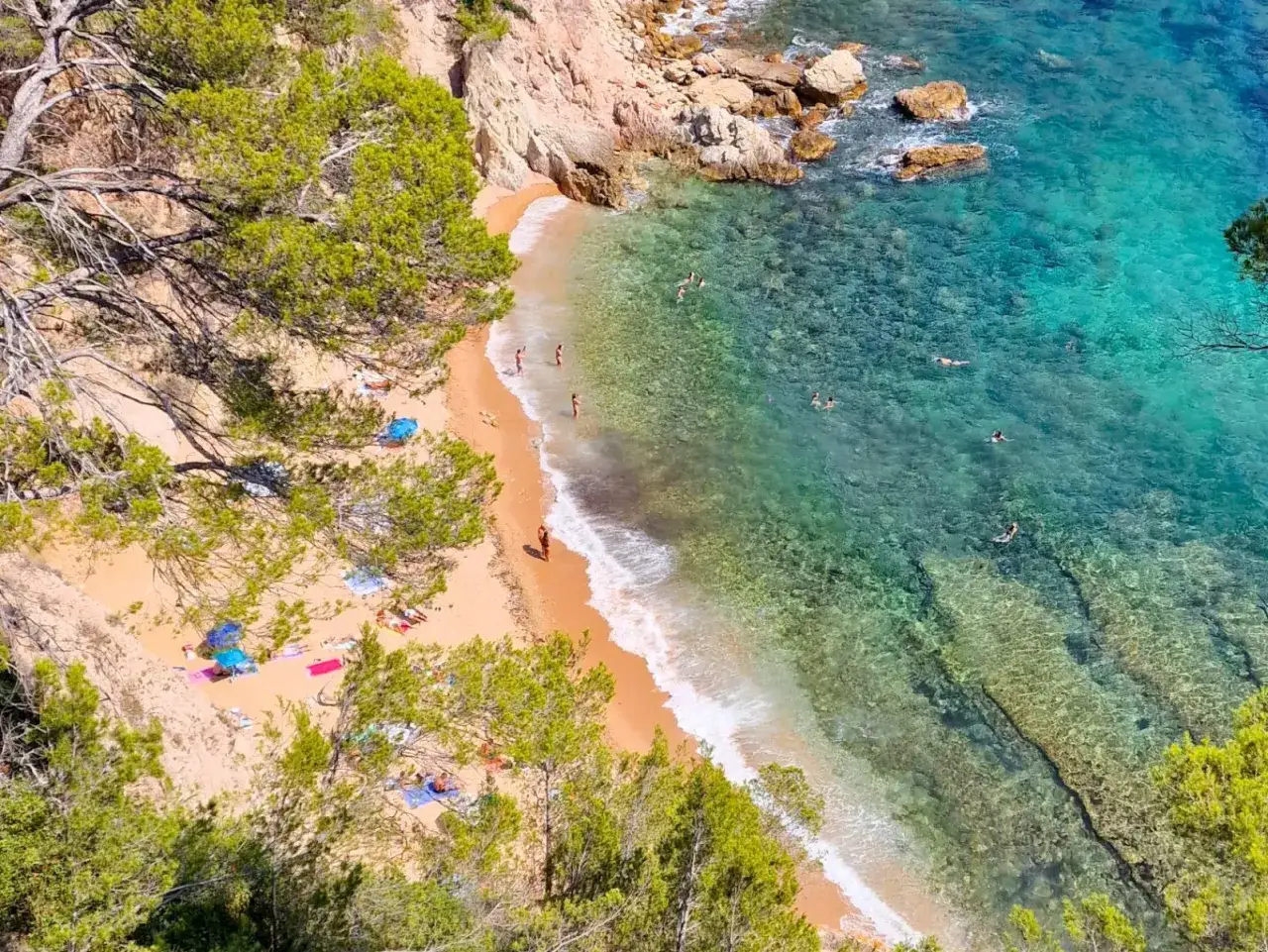 Una cala idílica con arena dorada, aguas cristalinas y gente disfrutando del mar. Los caminos de ronda ofrecen vistas espectaculares de este paraíso.