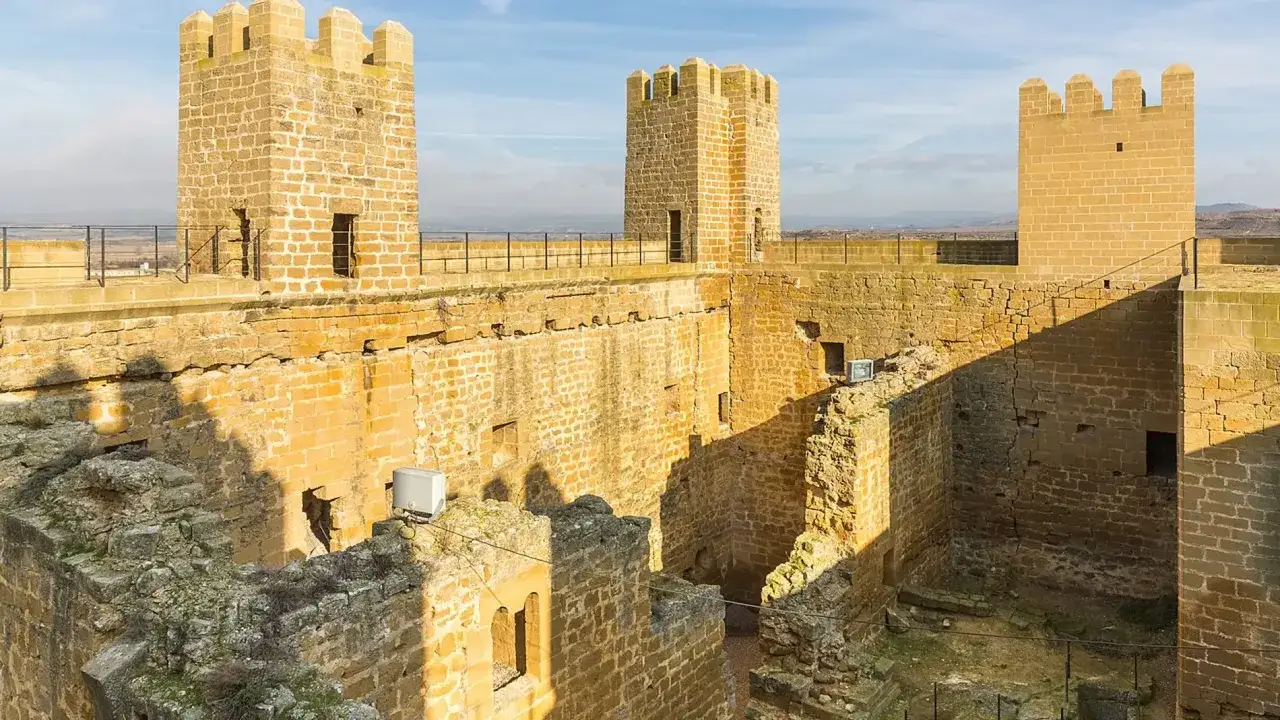 Torres almenadas y muros de piedra dorada del castillo de Sadaba, con un patio interior y el cielo azul de fondo.