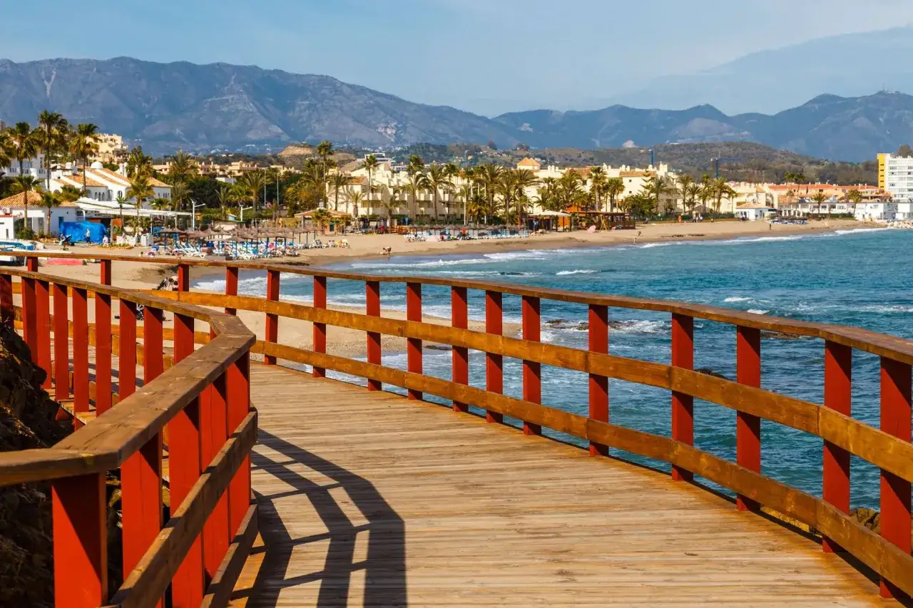 Paseo marítimo de madera con barandillas rojas y mar azul. Al fondo, la playa y edificios de Mijas Costa con montañas.