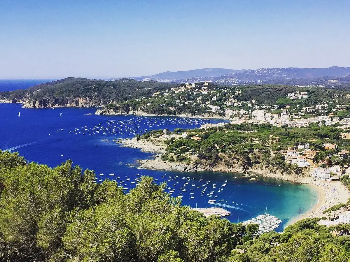 Vista panorámica de la costa con el faro de Sant Sebastià, numerosas embarcaciones en el mar azul y casas blancas entre pinos.