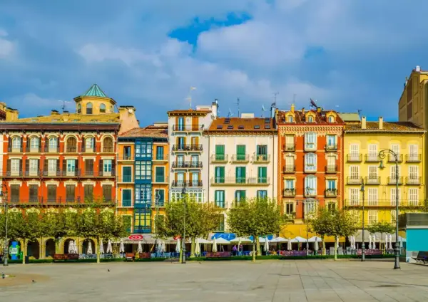 Fachada de edificios coloridos en una plaza, ideal para saber **qué ver en Pamplona**. Terrazas y árboles verdes bajo un cielo azul.