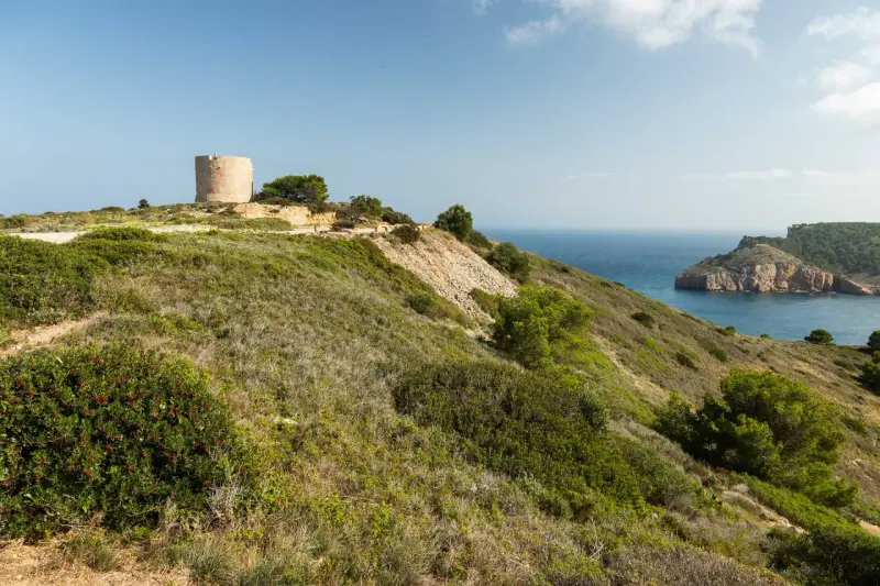 La torre de Montgó se alza sobre una colina verde, vigilando el mar azul y una bahía rocosa.