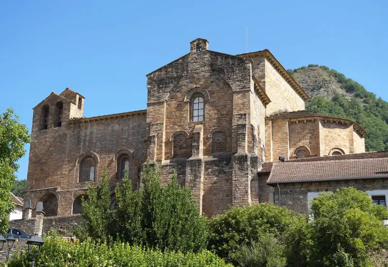El **monasterio de Siresa**, con su imponente arquitectura de piedra y campanario, se alza bajo un cielo azul, rodeado de vegetación exuberante.