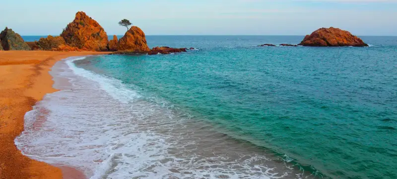 La platja mar menuda, con sus rocas anaranjadas y aguas turquesas, invita a soñar. Una ola rompe suavemente en la arena dorada.